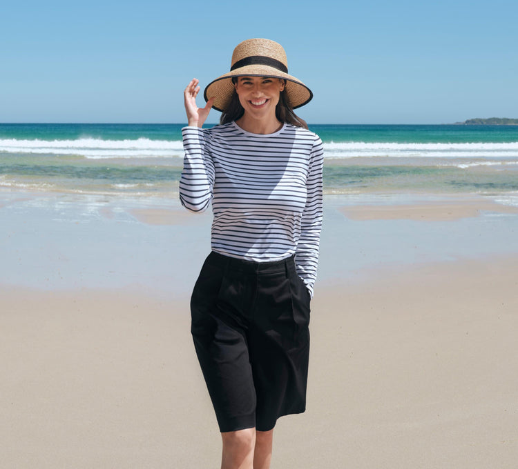 Woman in a Solbari striped shirt and wide-brimmed raffia hat standing on a beach with ocean in the background.
