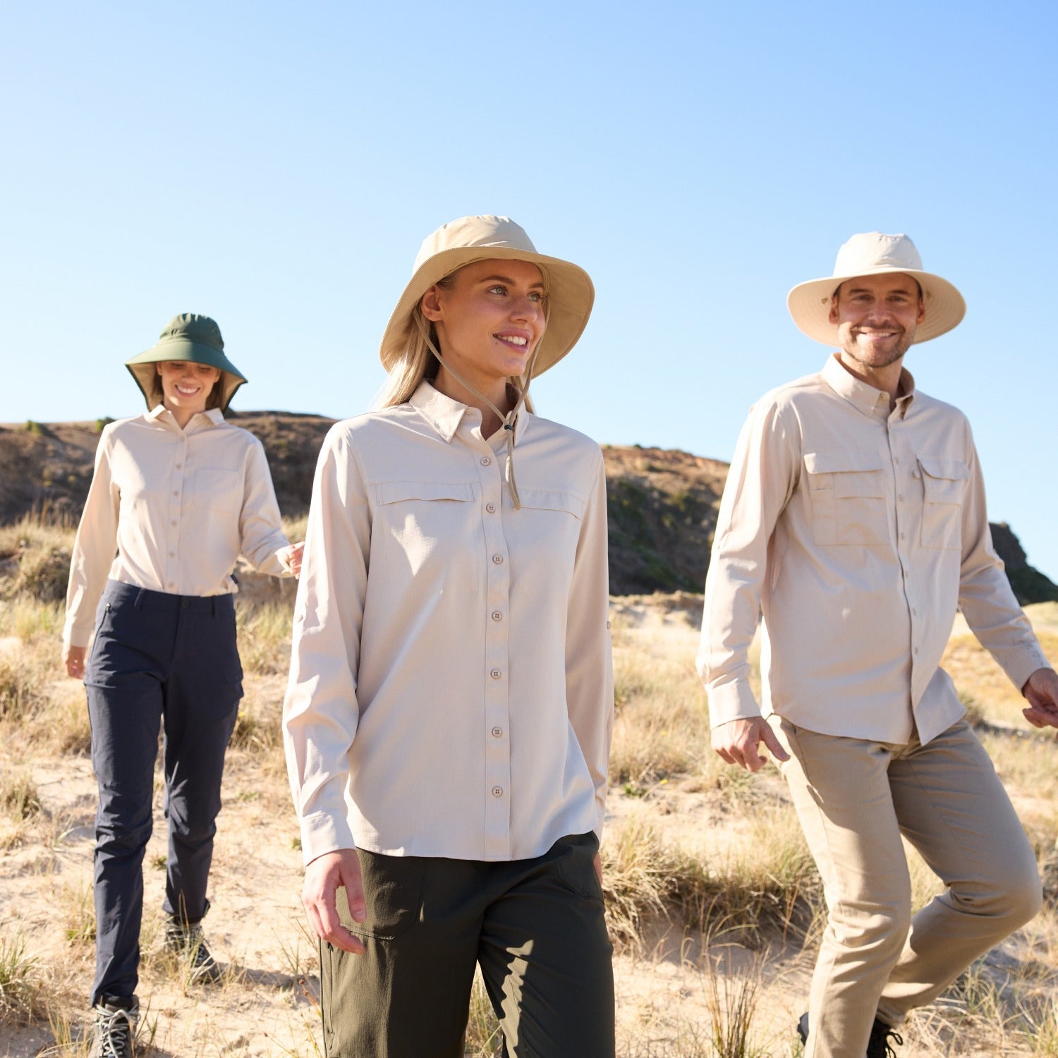 Three people walking outdoors in a desert-like environment wearing hats and light clothing.